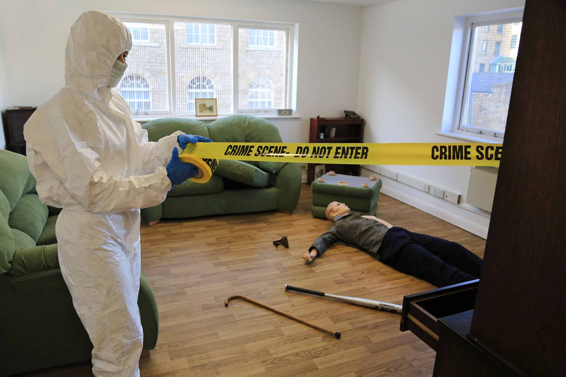 Someone in a forensics outfit is pulling a yellow ribbon around a mock crime scene. 