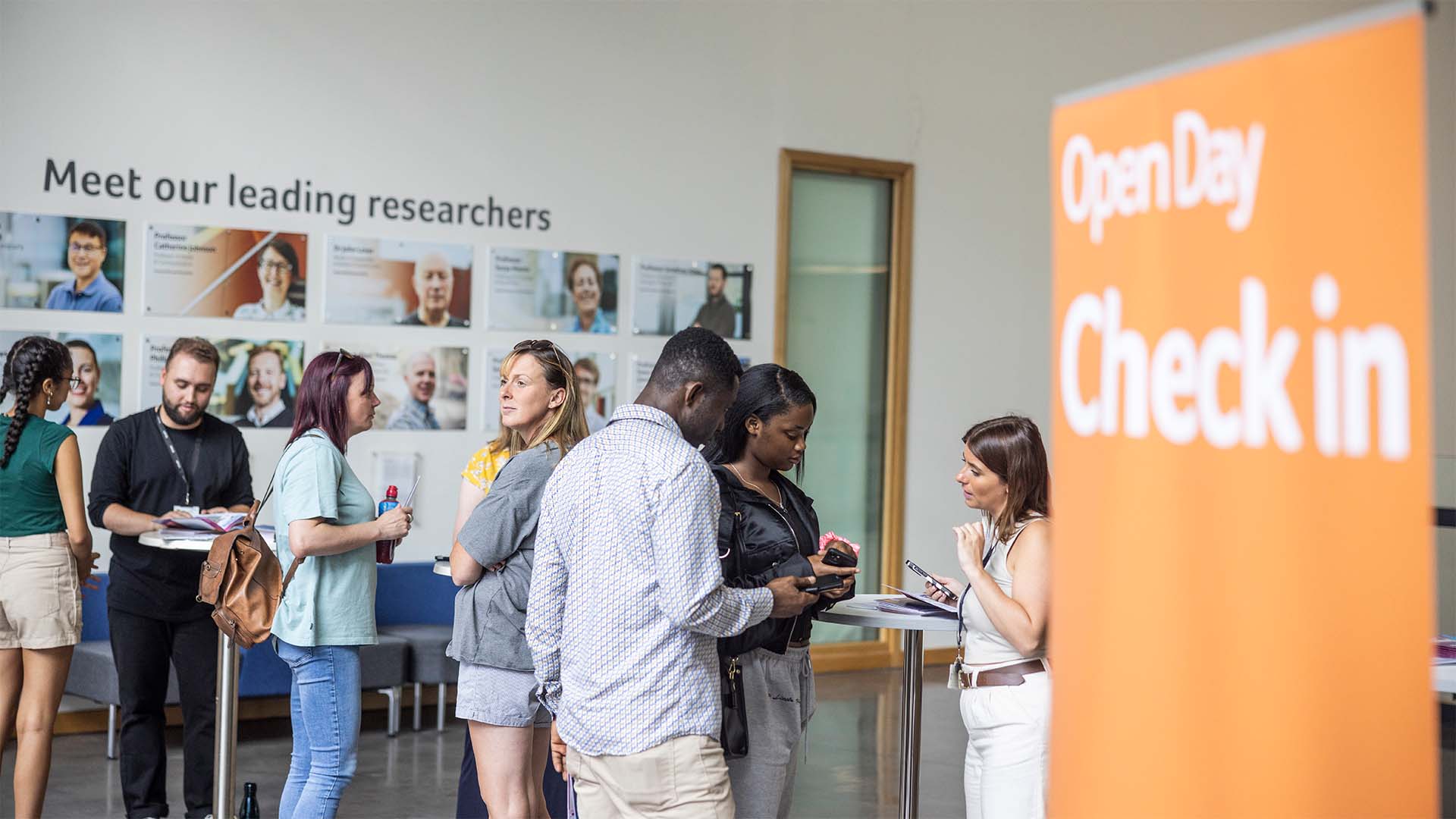Group of attendees checking in at an Open Day event near a 'Check-in' sign, with posters reading 'Meet our researchers' in the background.
