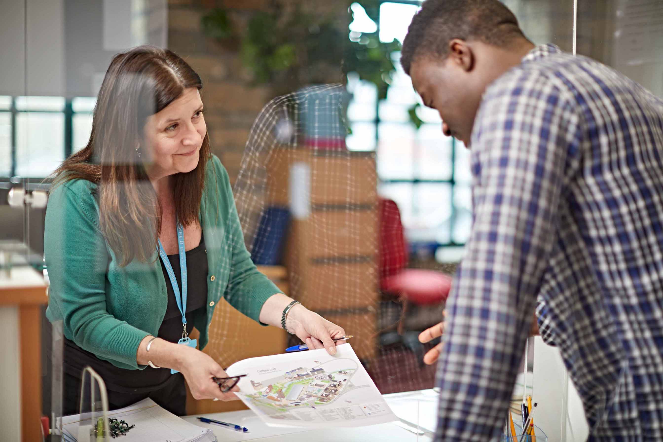 A woman with dark hair and a green top shows a map to a student wearing a check shirt