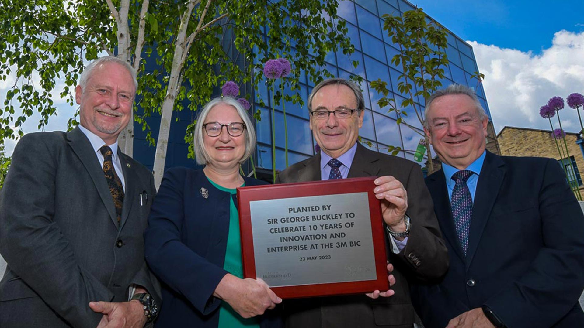 Four people infront of the 3M BIC building in Huddersfield with a plaque to mark the anniversary