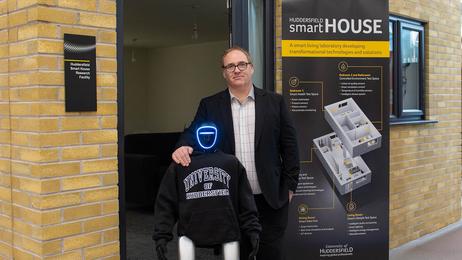 A man standing in the doorway of a house with a humanoid robot wearing a hoodie