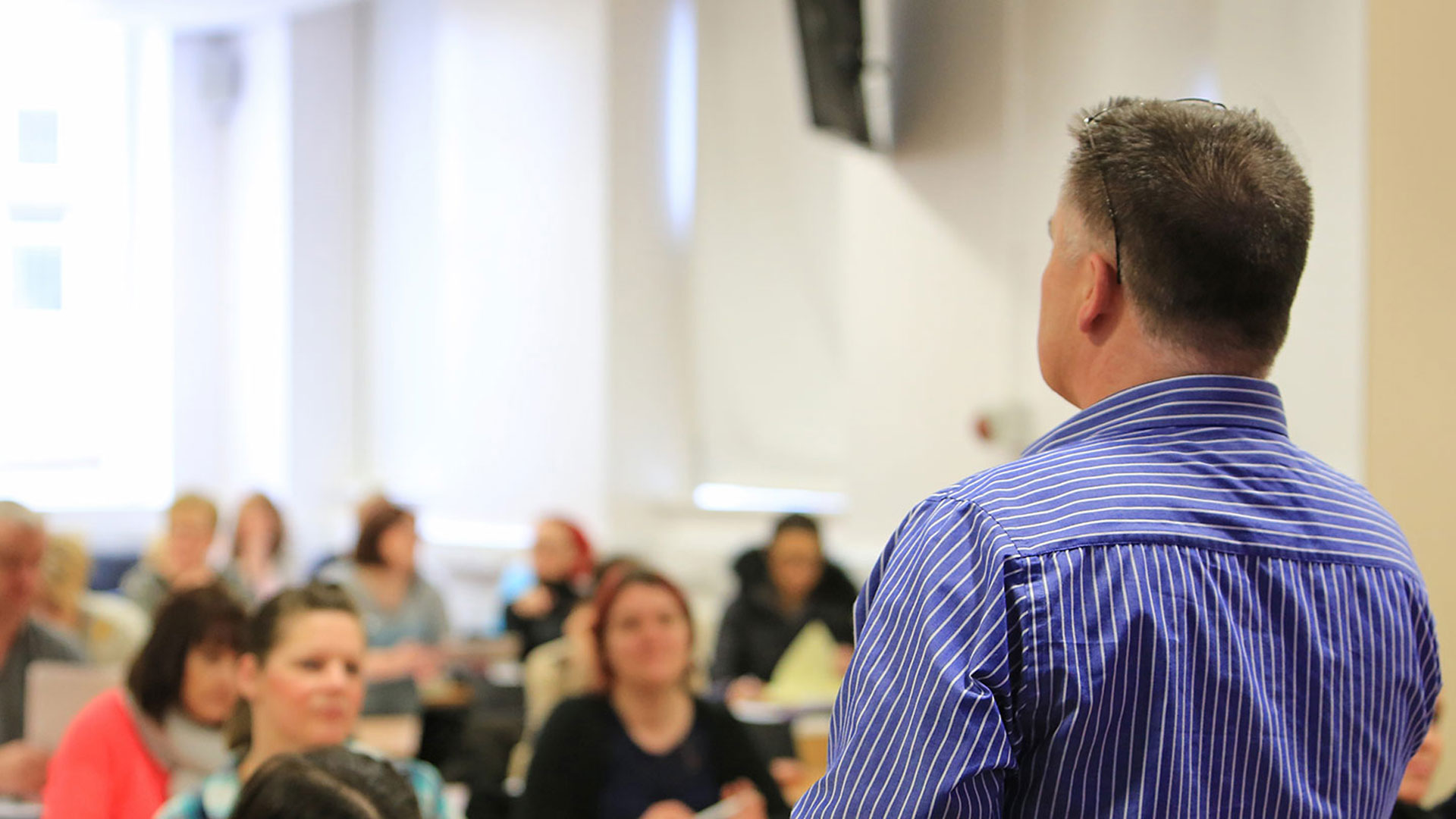 A teacher with blue striped shirt faces a classroom of students. Students faces are blurred out. 